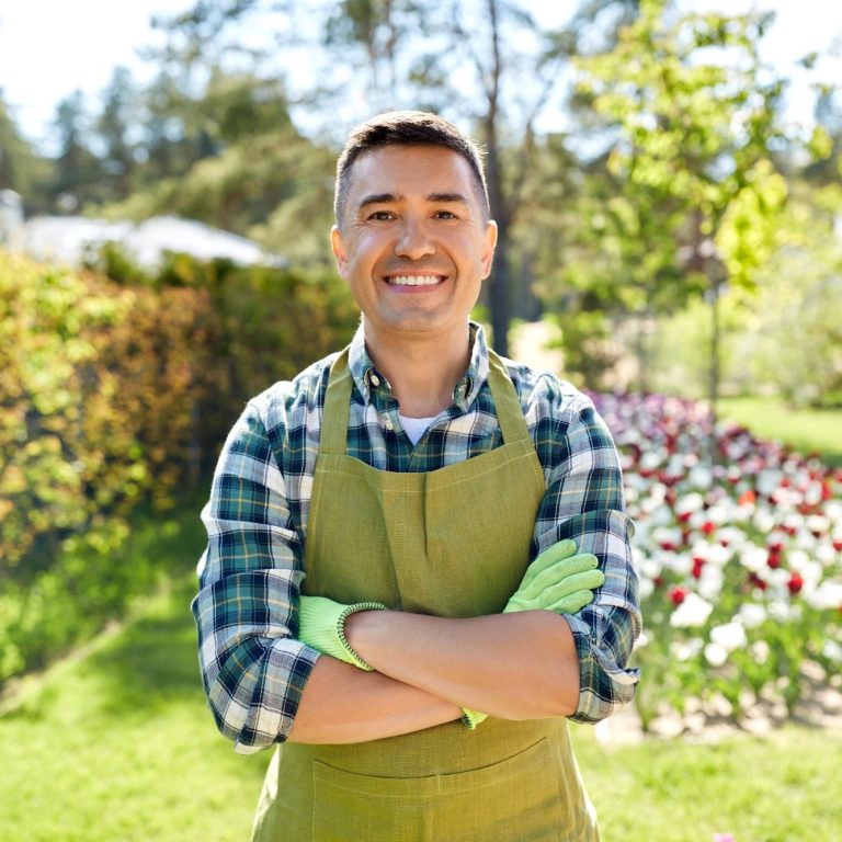 Mann im Garten mit grünem Schürze und Handschuhen, lächelt vor bunten Blumen.