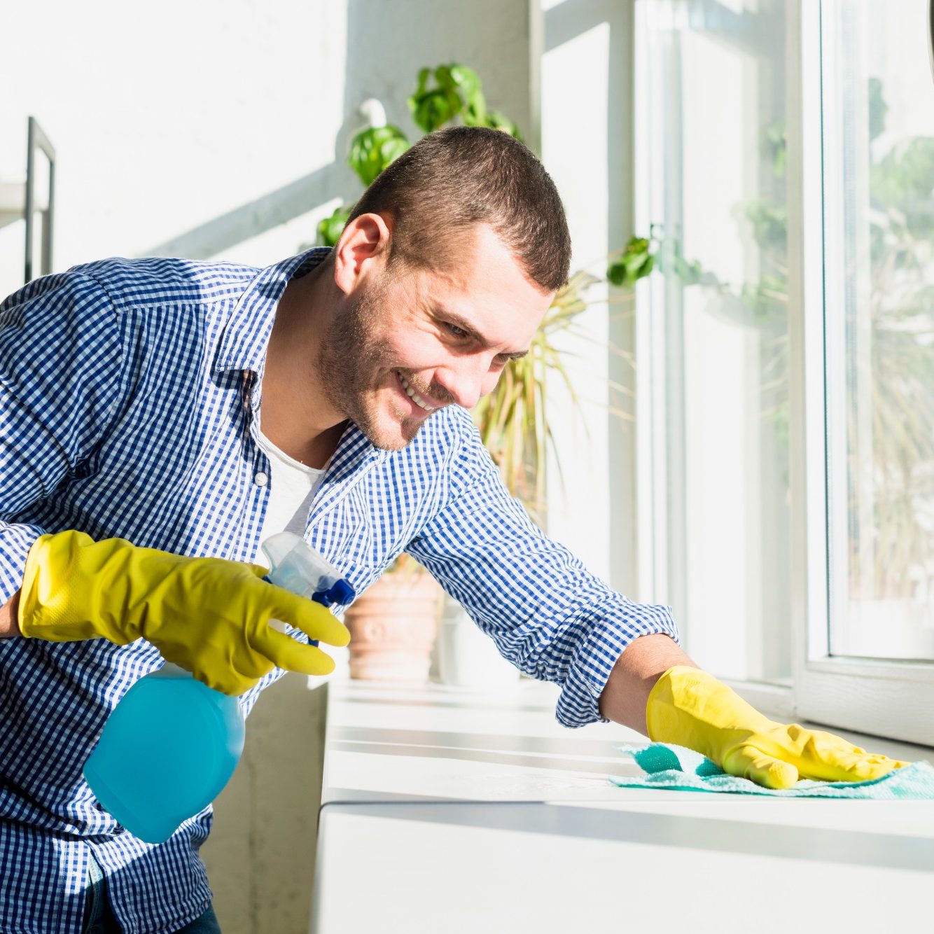 Mann mit gelben Handschuhen reinigt das Fensterbrett mit einem Tuch und Spray.