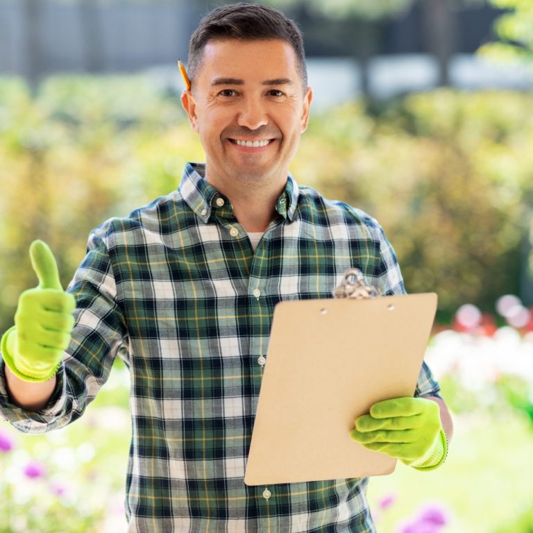 Mann im Garten mit grünem Handwerkshandschuh und Clipboard, zeigt Daumen hoch.