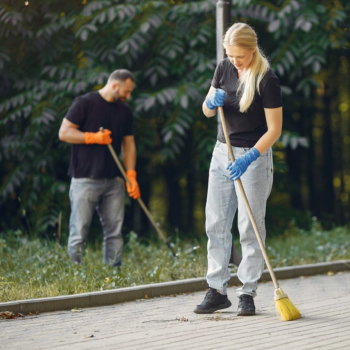 Zwei Personen reinigen einen Parkweg mit Besen und Handschuhen.